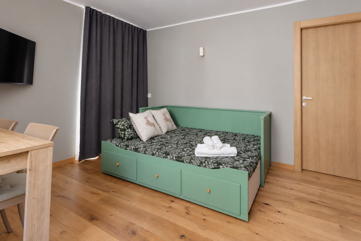 Een kamer met een groen slaapbank, een houten eettafel, een aan de muur bevestigde televisie en een houten deur.