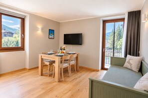 A room with a dining table set for four, a flat-screen TV mounted on the wall, a sofa with a cushion, and a window with a view of trees and mountains. - Vrbo Property (Cepina)