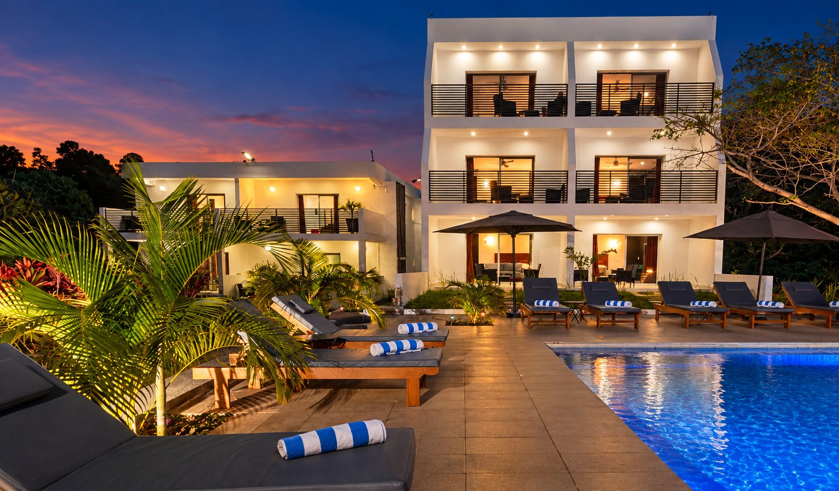 A modern two-story building with a swimming pool, sun loungers, and outdoor lighting.