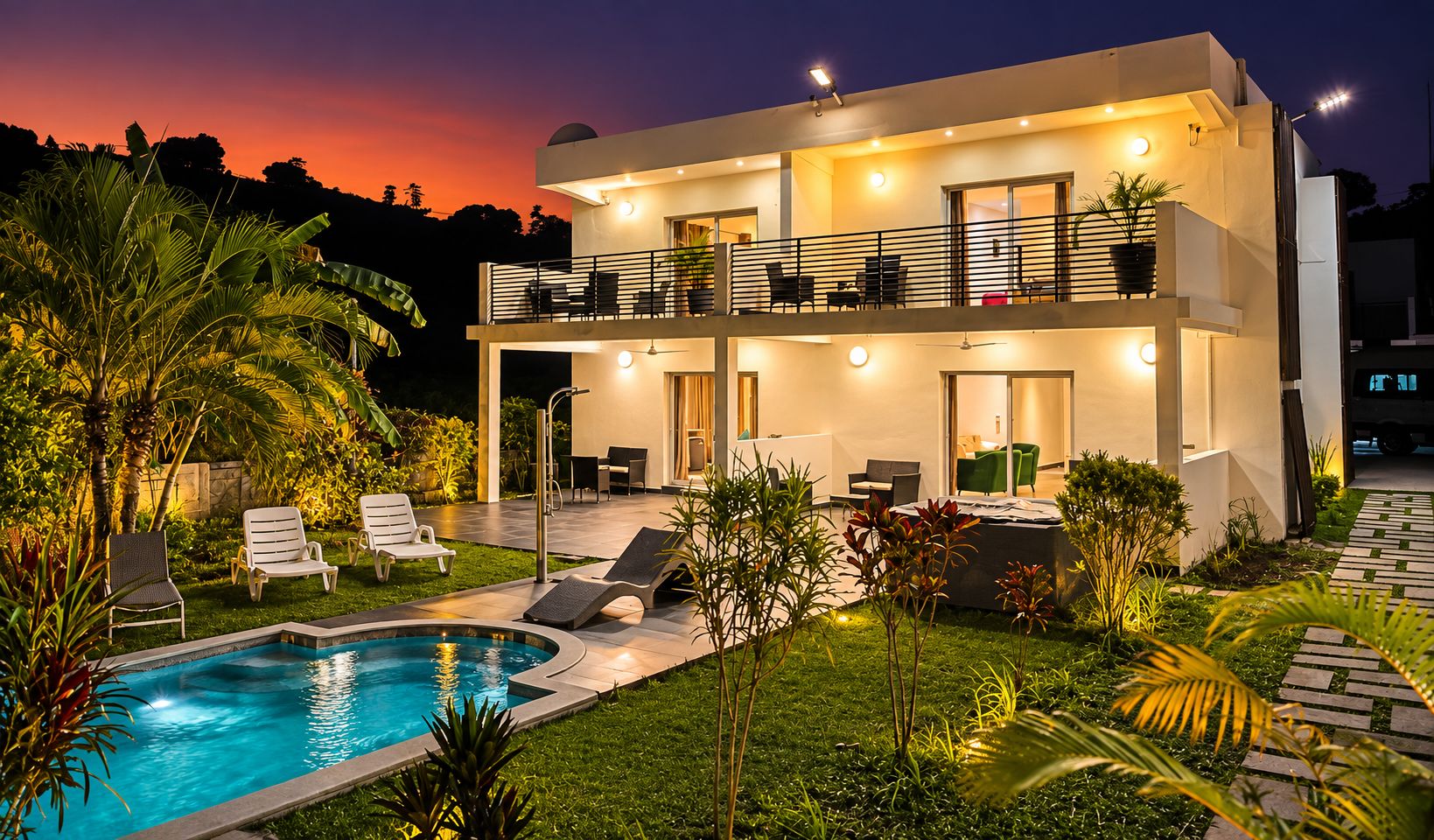 Una casa moderna de dos pisos con piscina, mobiliario exterior e iluminado balcón.