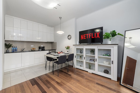 Une cuisine moderne avec des meubles blancs, un coin repas avec une table et des chaises, un téléviseur posé sur un meuble et une horloge au mur.