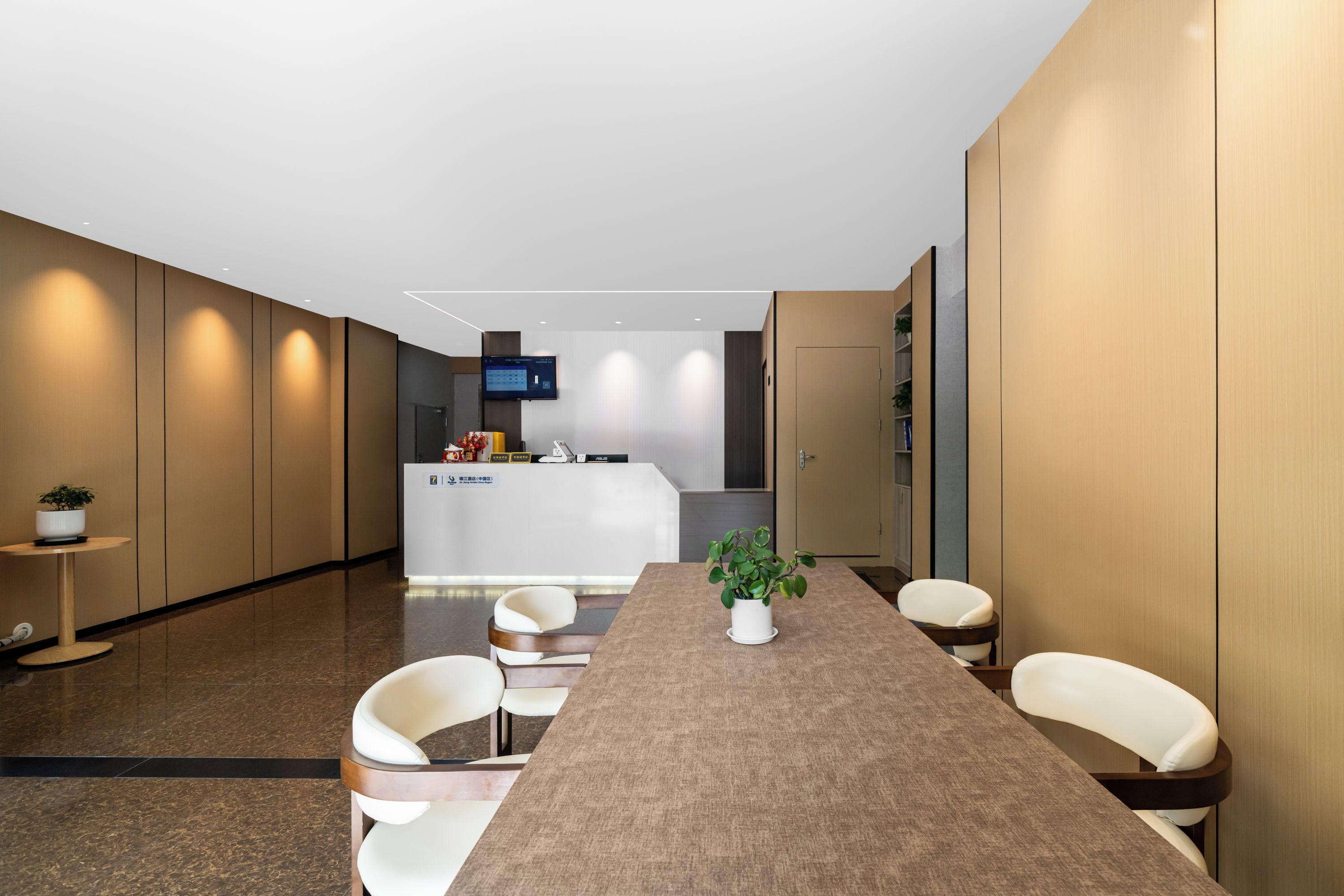 A modern hotel lobby with a long reception desk, seating area, and a kitchen in the background.