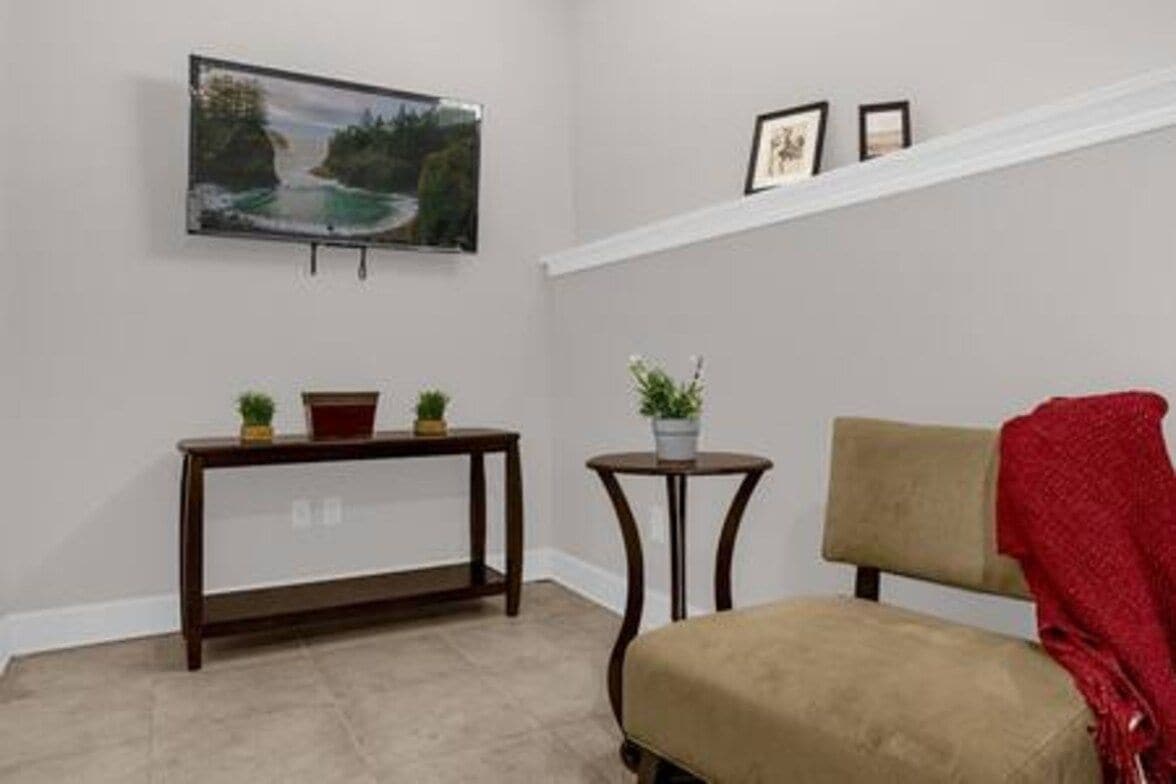Een kamer met een beige stoel, een klein rond tafeltje met een plant in pot, een groter houten tafeltje met twee planten in pot, een ingelijst beeld aan de muur en een trap met een witte leuning.