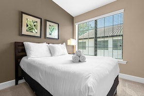 A bedroom with a large bed, two framed botanical prints on the wall, a window with blinds, and a lamp on the nightstand. - Family Vacation Retreat With Resort Pool Games (Orlando)