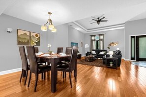 Un salon moderne avec un coin repas, doté d’un ventilateur de plafond, d’un plancher en bois et d’une grande fenêtre avec des rideaux.