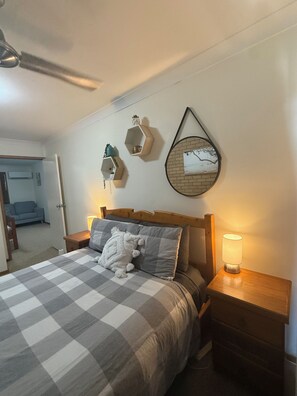 Ein Schlafzimmer mit kariertem Überwurf, einem Holzkopfteil, zwei Nachttischen mit Lampen sowie Wanddekorationen, darunter hexagonale Regale und ein runder Spiegel.