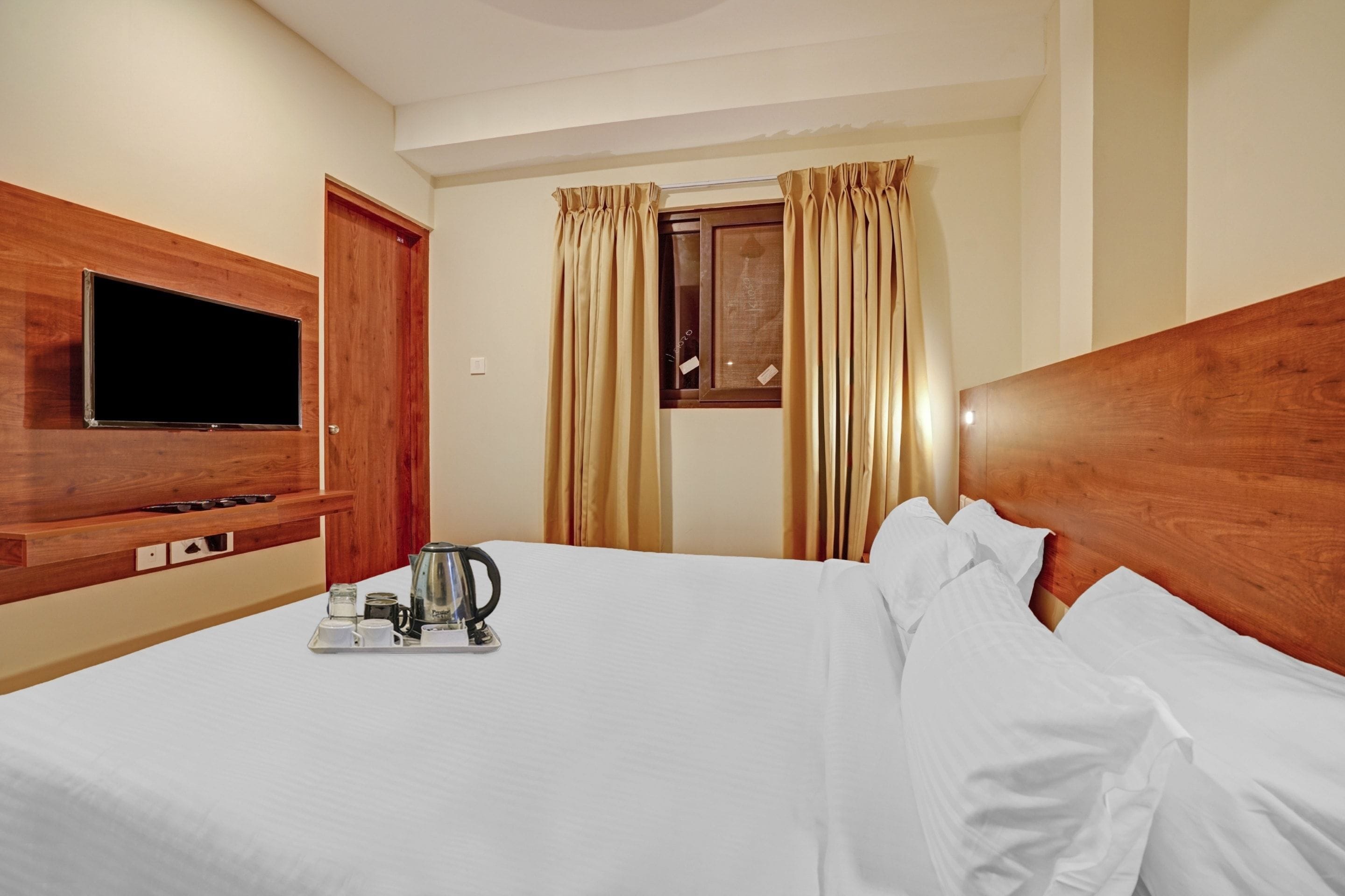Une chambre d’hôtel avec un lit, un téléviseur fixé au mur, une fenêtre avec des rideaux et un plateau avec une bouilloire et des tasses.