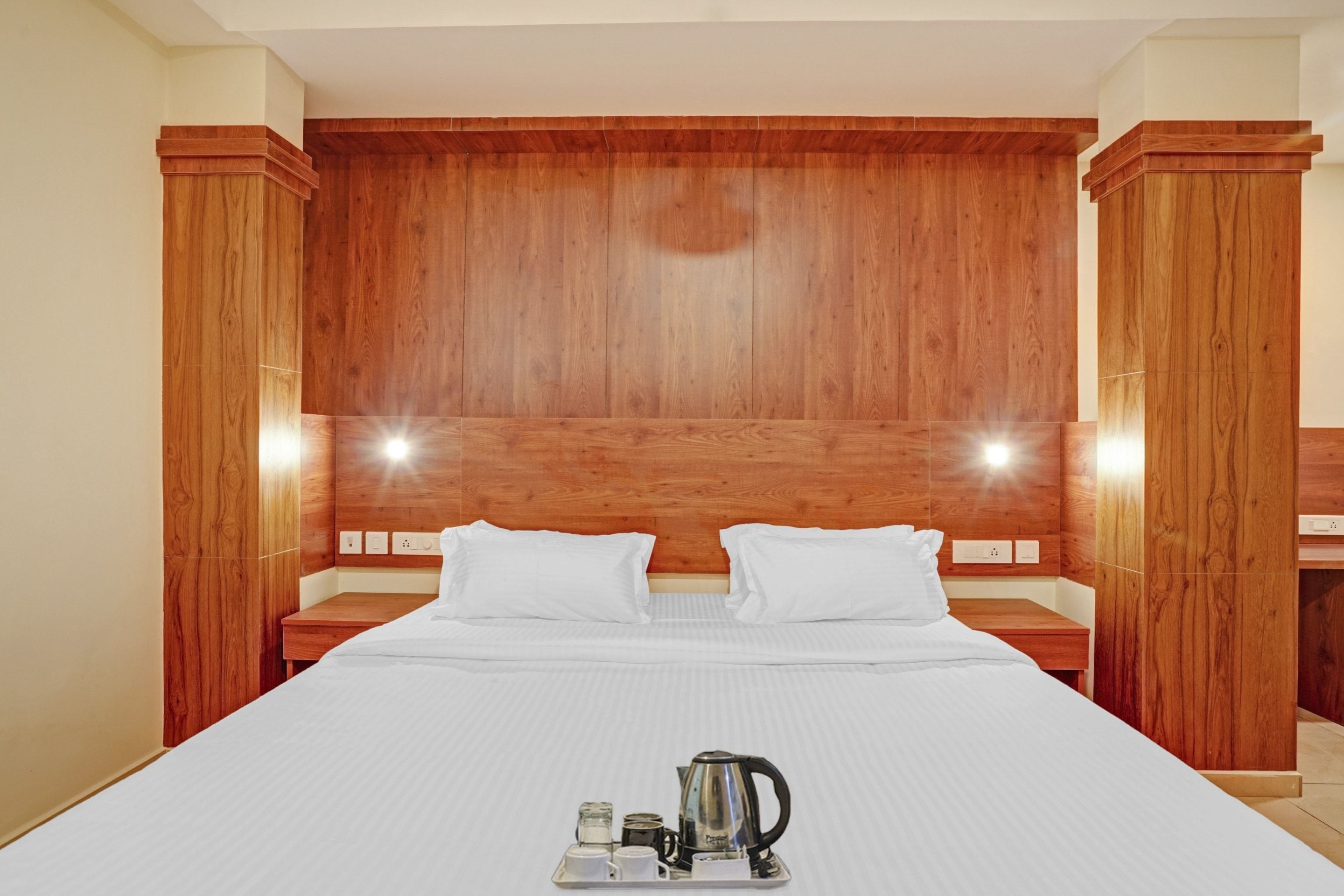 A hotel room with a wooden headboard, two bedside tables, a bed with white bedding, and a tray with a kettle and cups.