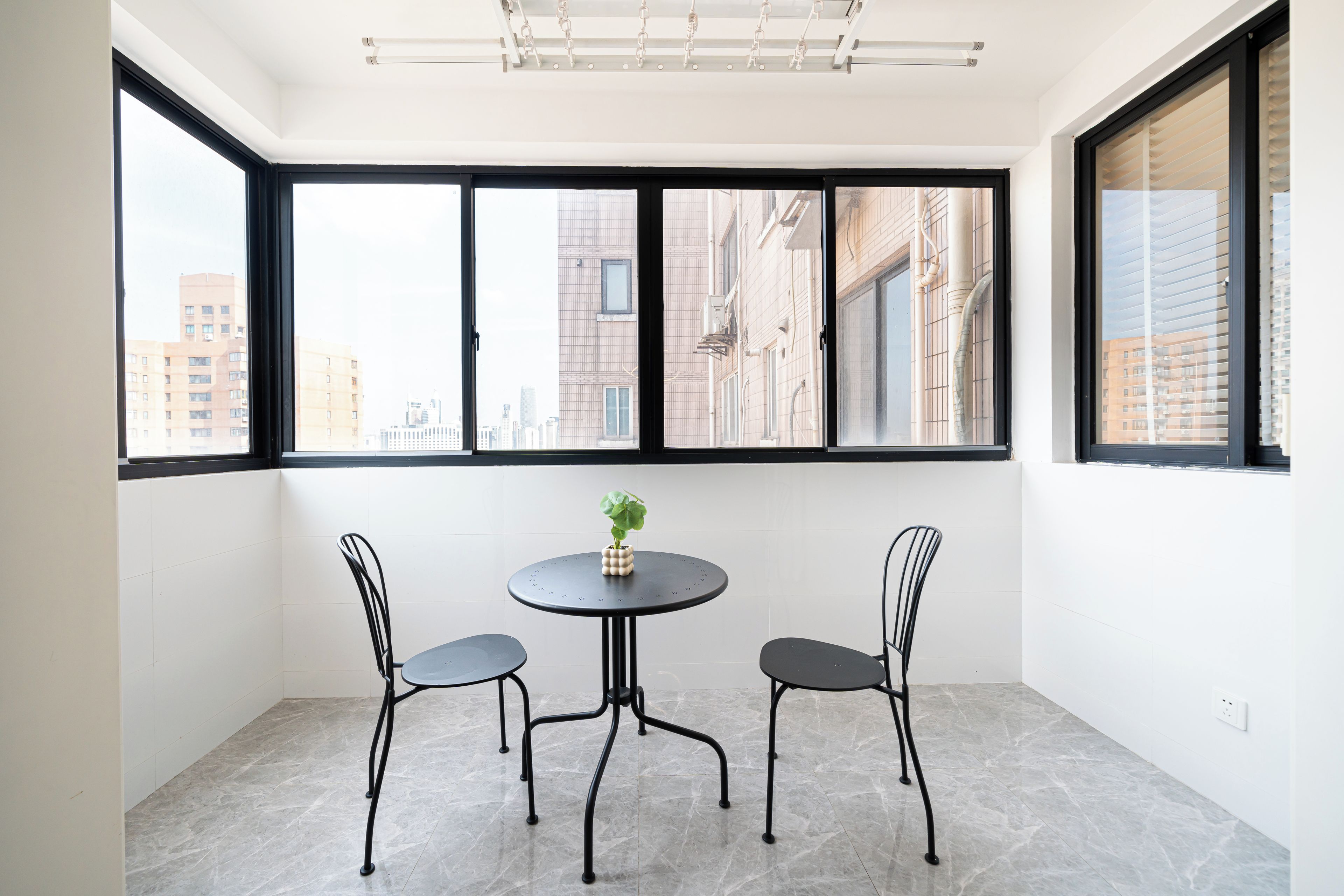 A small room with a round table and two chairs, large windows with a city view, and a ceiling light fixture.