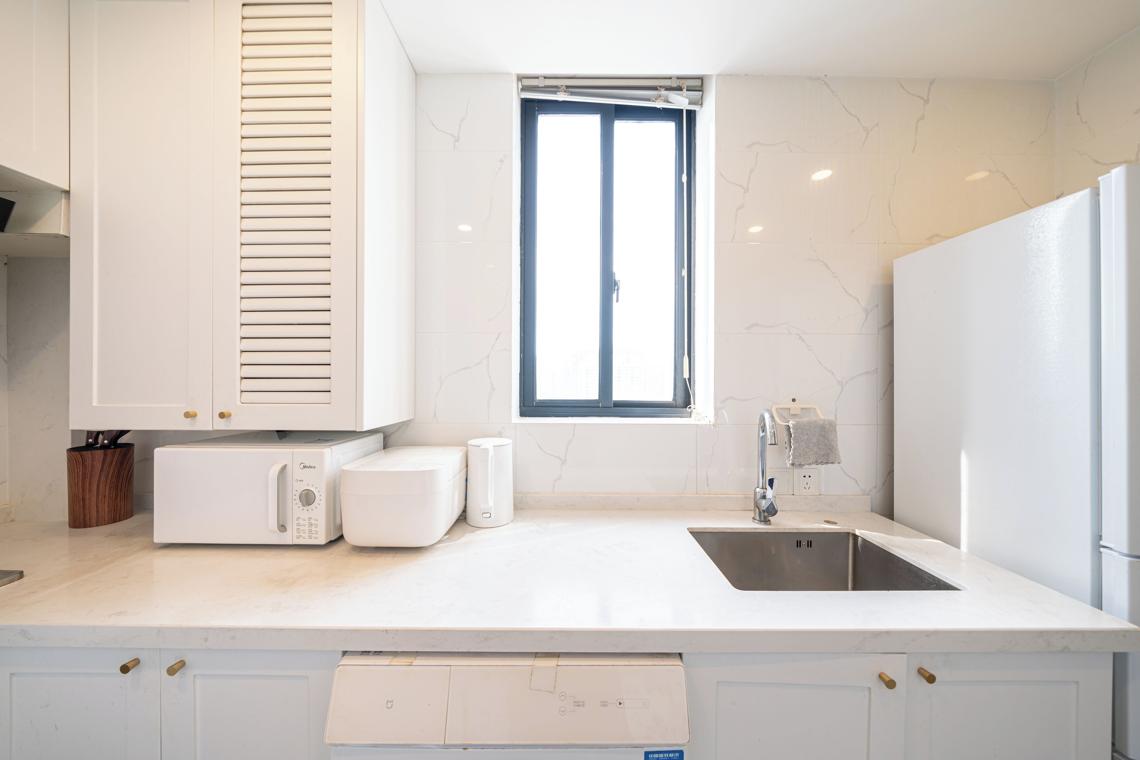Una cocina moderna con armarios blancos, microondas integrado, un lavamanos y una ventana con persianas.