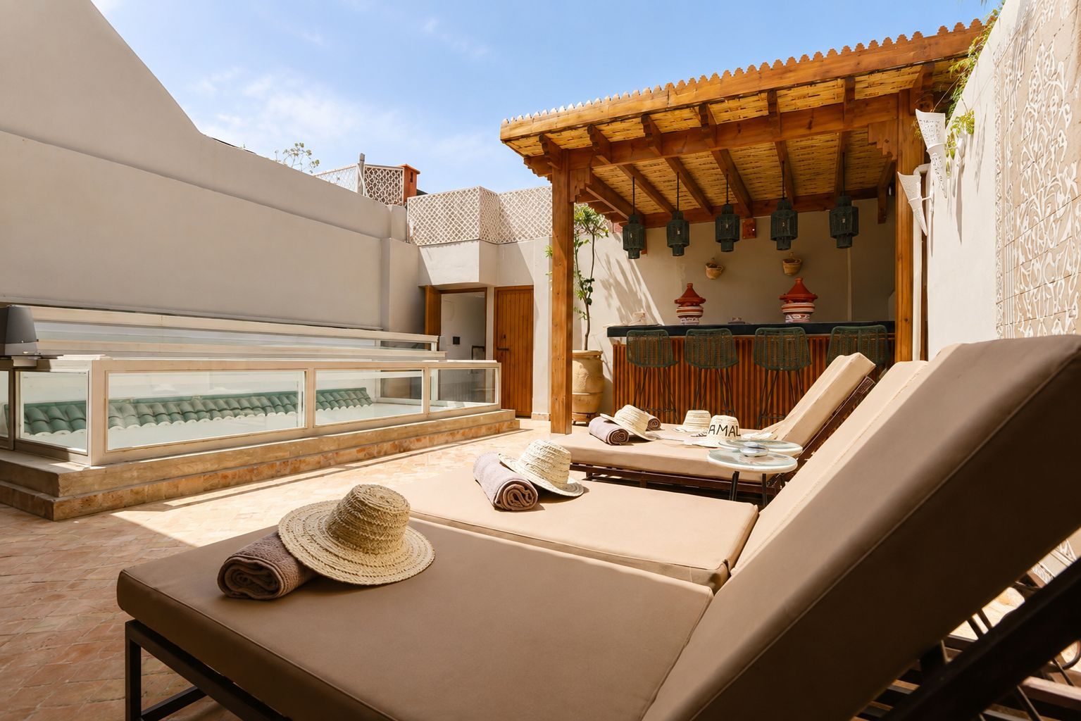 A sunlit outdoor lounge area with lounge chairs, a glass-enclosed pool, and a wooden pergola.