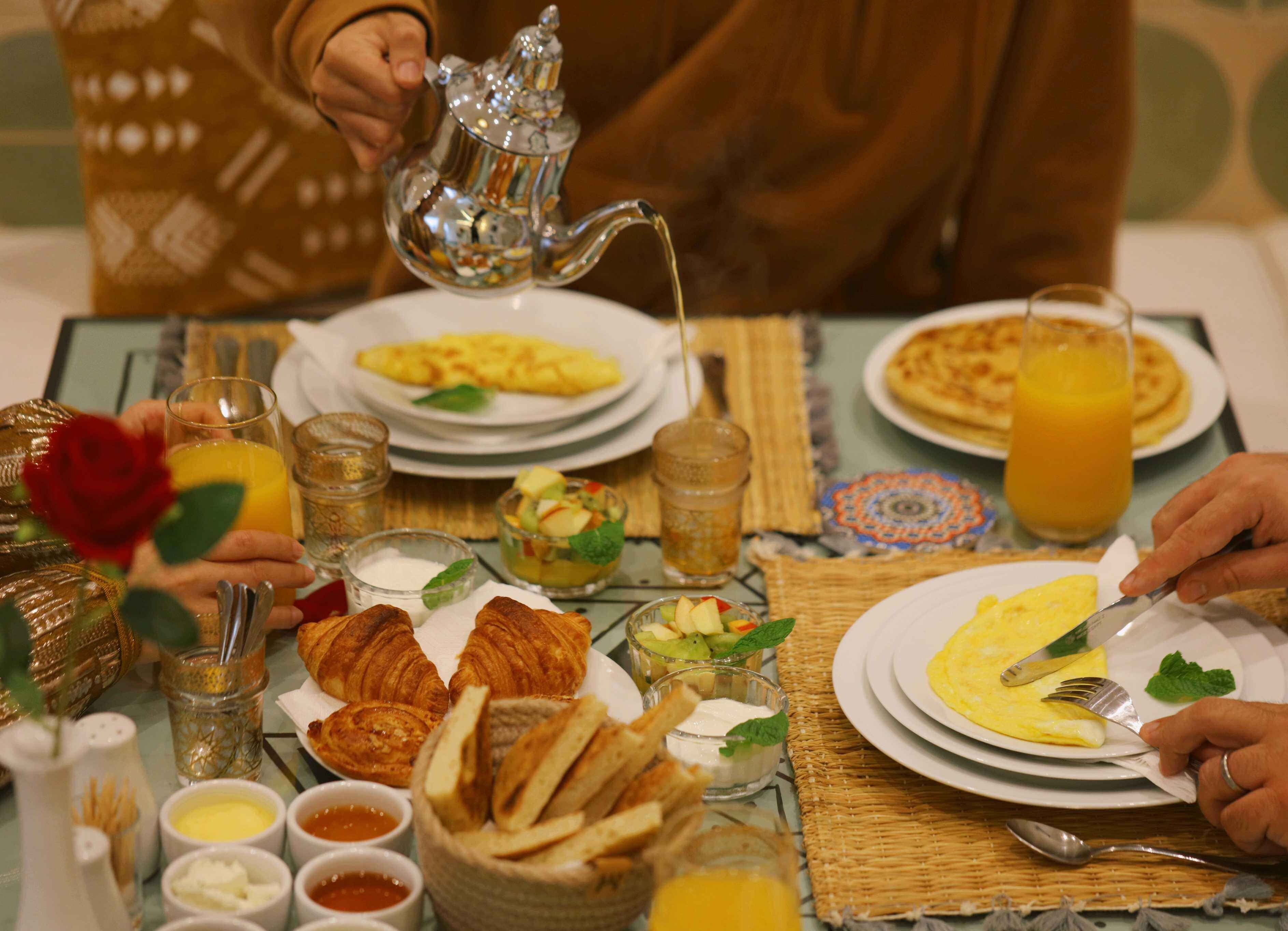 Una escena de desayuno con platos de huevos, tostadas y fruta, junto con diversos condimentos y una tetera.