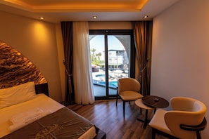 A hotel room with a bed, two chairs, a small table, and a view of a pool and buildings outside. - GOLD SANDORA HOTEL (ARSUZ)