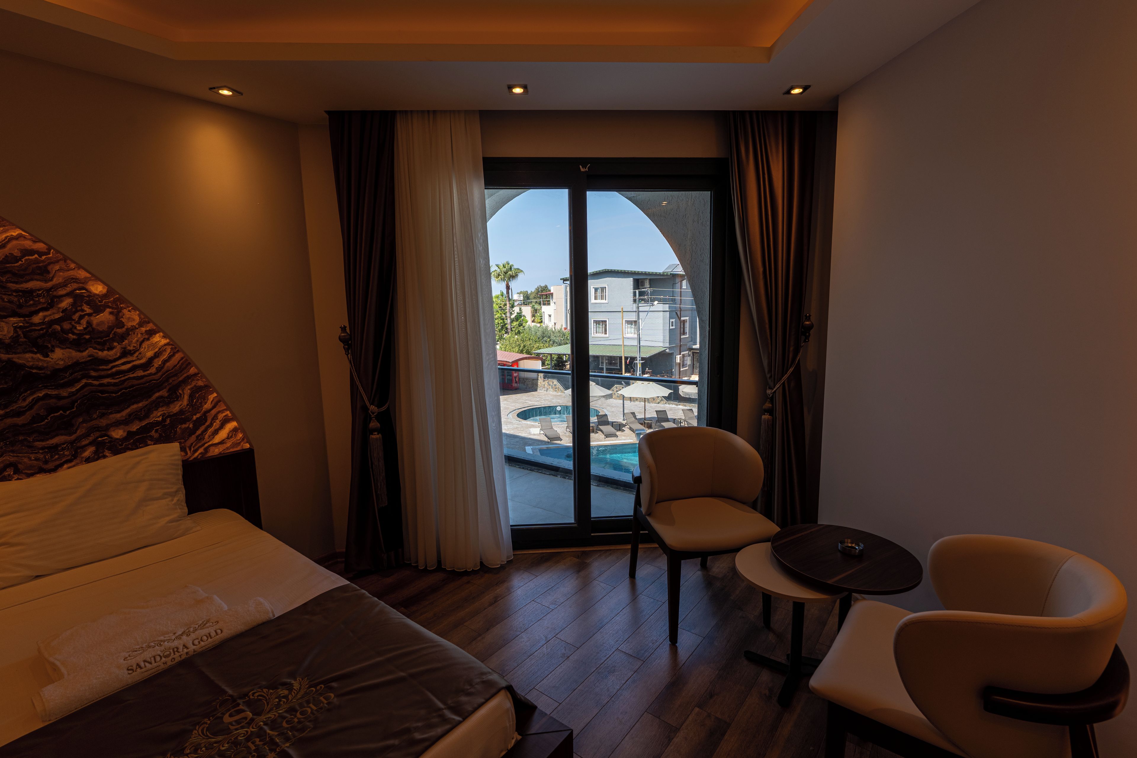 Habitación de hotel con cama, dos sillas, una mesita y vistas a una piscina y edificios.