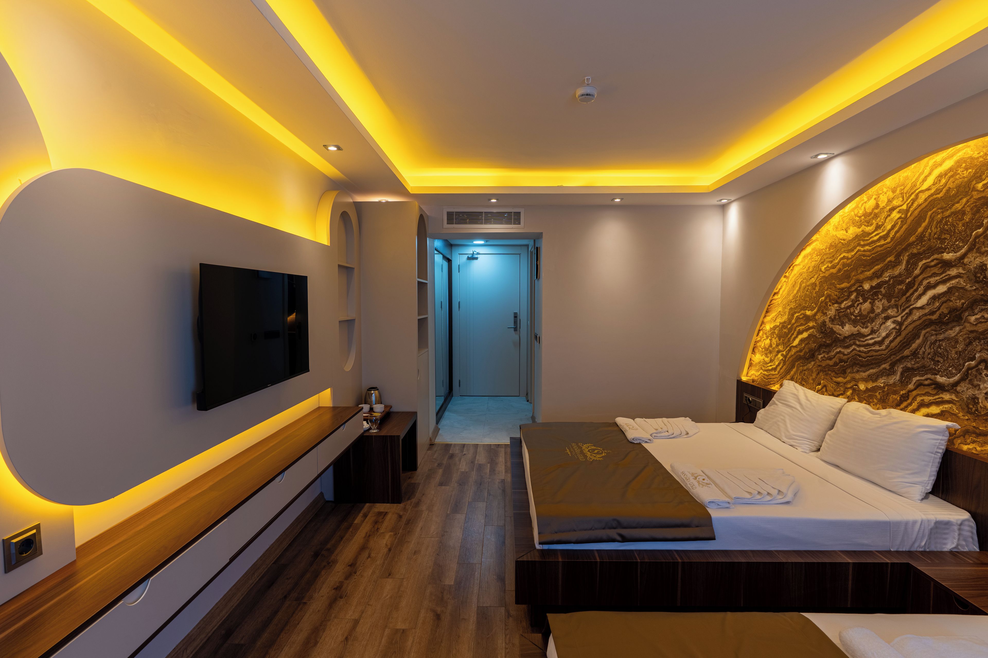 Una habitación de hotel moderna con una televisión de pantalla plana, una cama con cabecera estampada en dorado, una mesita de noche de madera y una pequeña mesa con un hervidor y tazas.
