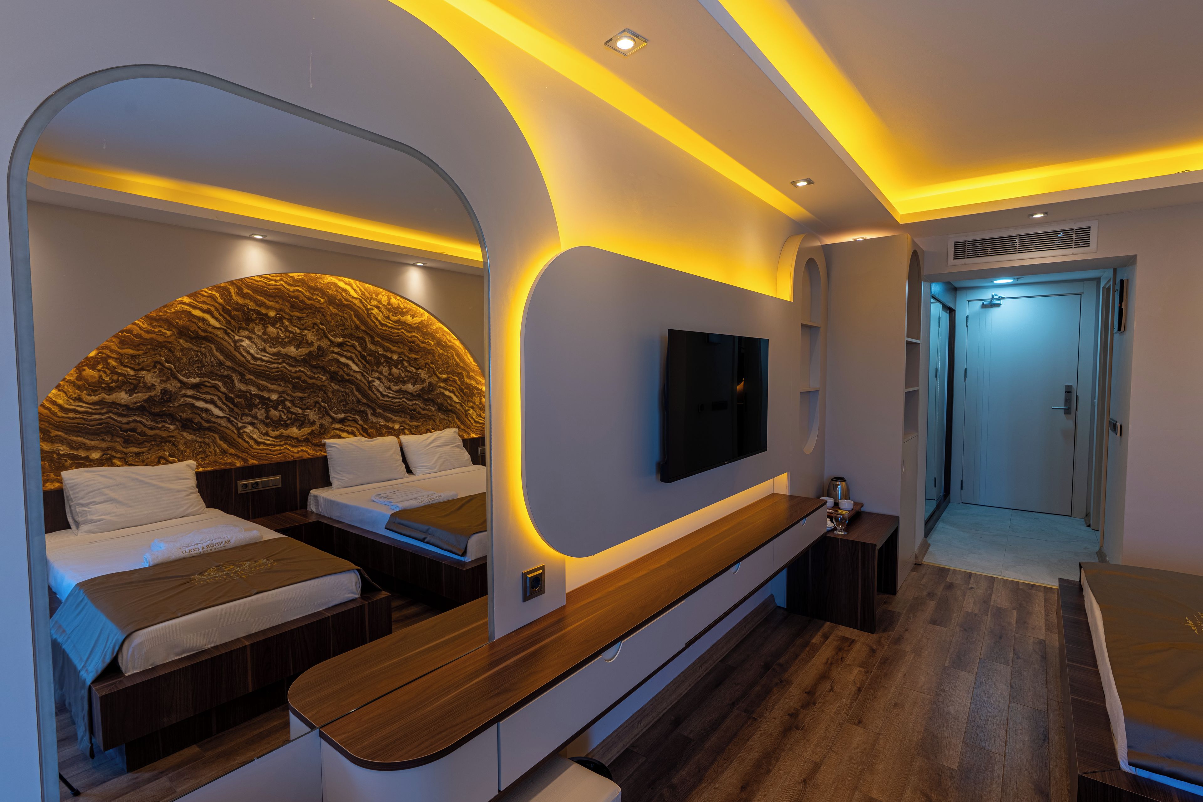 A modern bedroom with a curved headboard, a flat-screen TV, a wooden bench, and a walk-in closet.