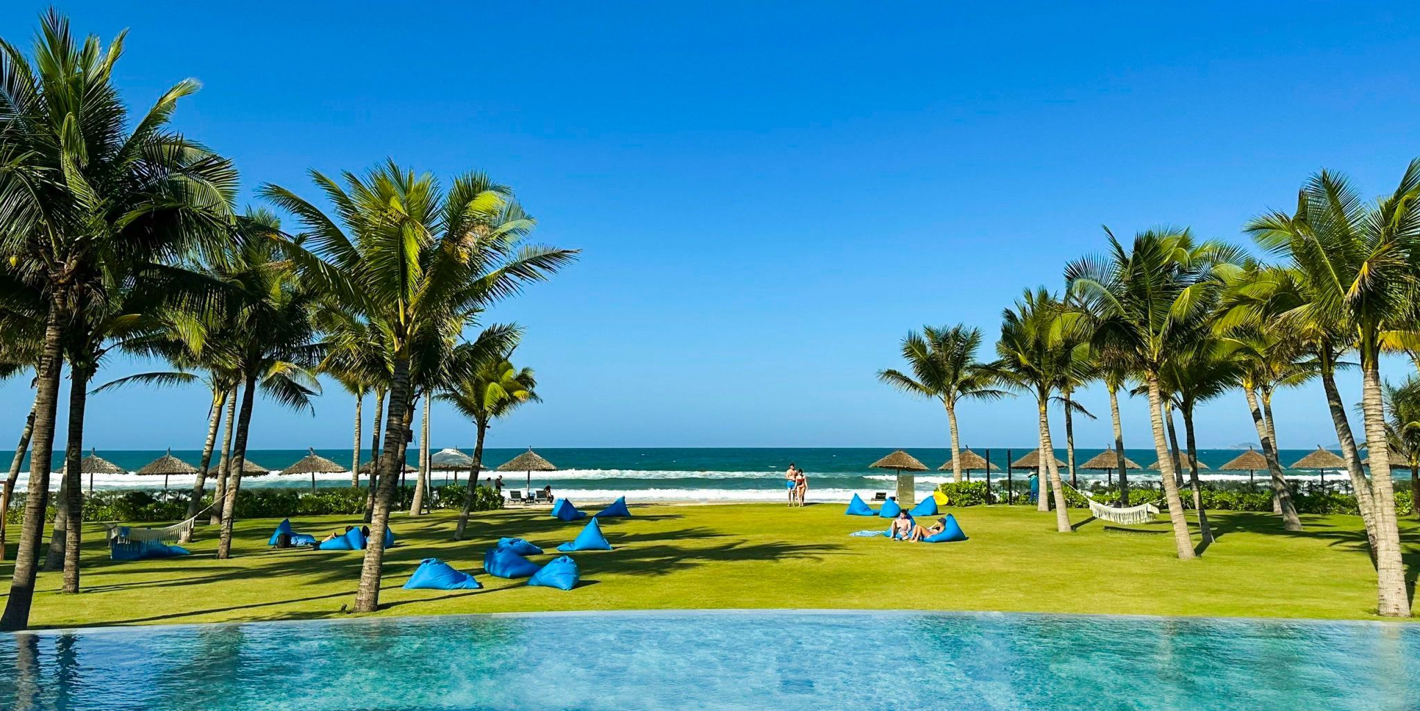 Zona junto a la piscina con sillones de descanso y sombrillas, palmeras y vistas al océano.