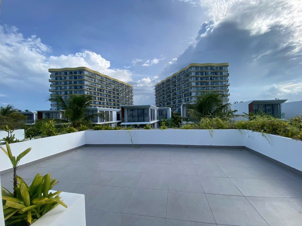 A rooftop terrace with a view of two high-rise buildings, surrounded by greenery and white walls.