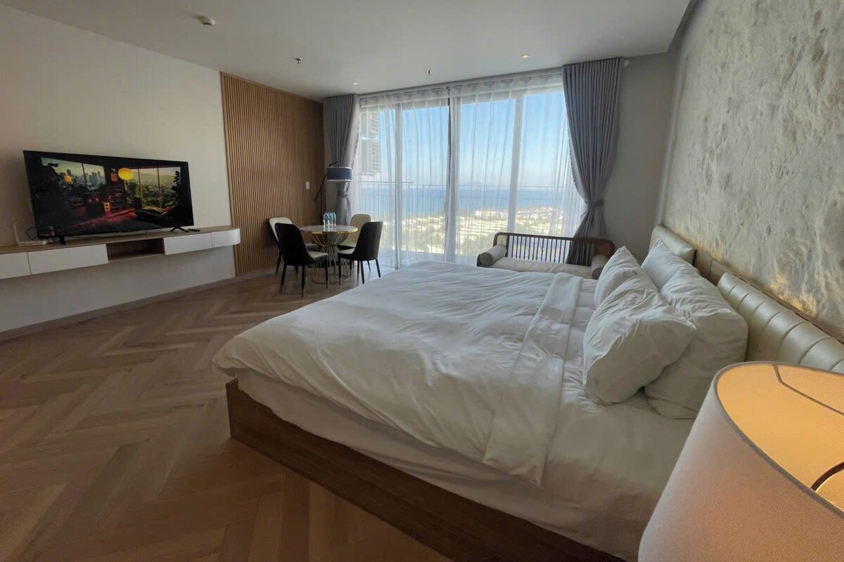 Habitación de hotel con una cama grande, televisor, mesa de comedor con sillas y vistas a la ciudad a través de amplios ventanales.