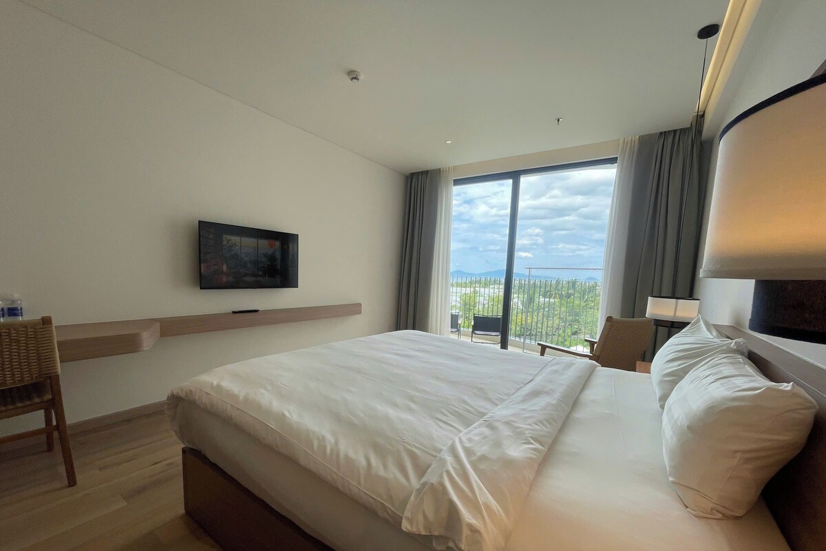 A hotel room with a large bed, a flat-screen TV mounted on the wall, a balcony with a view, and a chair.