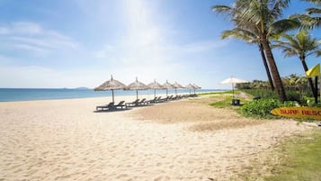 A beach with sun loungers and umbrellas, a sign indicating a surf rescue area, and palm trees.
