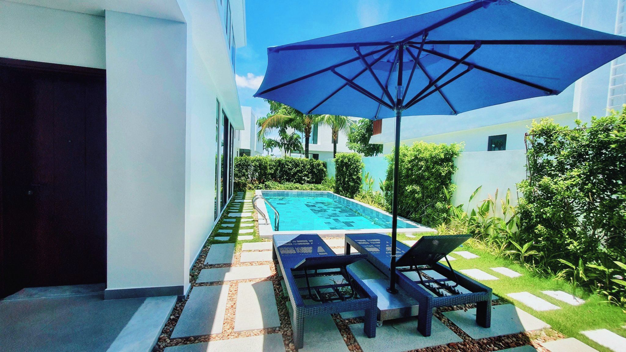 Zona de piscina con una sombrilla azul, sillas de descanso y un paseo de baldosas.