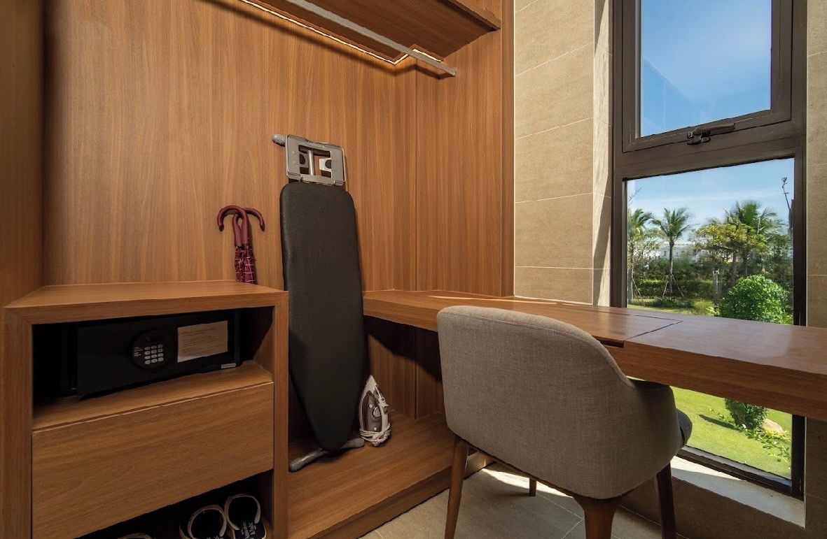 Een kamer met een houten bureau, een stoel, een raam met uitzicht op groen en een wandplank met een strijkplank.