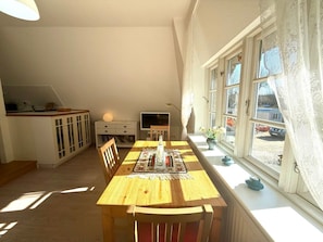 A dining area with a wooden table and chairs, a television, and a window with curtains. - "reetstubchen" - House Shell (Nieblum)