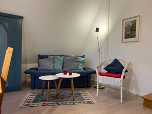 A room with a blue sofa, two small round tables, a white wicker chair, a patterned rug, and a painting on the wall. - "reetstubchen" - House Shell (Nieblum)