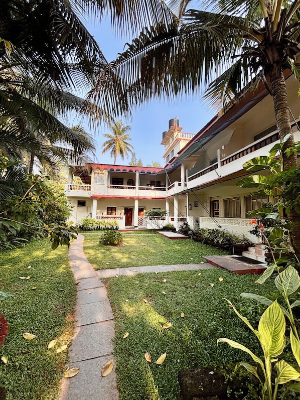 A courtyard with a pathway leading to a building with a red roof, surrounded by palm trees and greenery. - Dmellos Seaview Home (Candolim)
