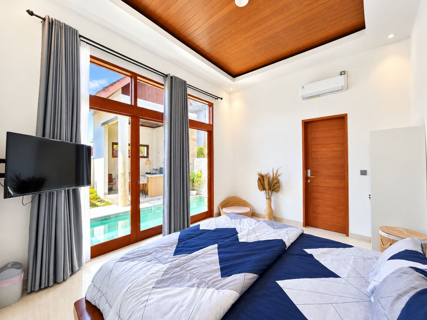 A bedroom with a large bed, a television, a sliding glass door to a balcony, and a wooden ceiling.