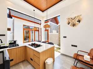 Eine moderne Küche mit Holzschränken, einer weißen Arbeitsplatte und einem eingebauten Herd. Es gibt einen Essbereich mit Tisch und Stühlen sowie eine kleine Sitzecke mit einem Sessel.