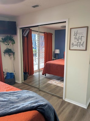 Ein Schlafzimmer mit einer Schiebetür zu einem Balkon, einem Bett mit orangefarbener Bettdecke, einem Nachttisch mit Lampe und einem motivierenden Zitat an der Wand.