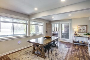 A dining area with a wooden table and chairs, a rug, and a view of a lake through large windows.