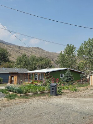 A green house with a porch, surrounded by a gravel driveway and a garden with flowers. - Cozy 2-Bdrm Pan Abode Home (Cache Creek)