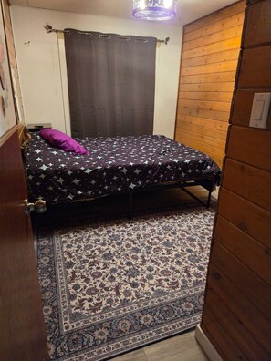 A small, single-bed room with a patterned bedspread, a purple pillow, and a decorative rug. - Cozy 2-Bdrm Pan Abode Home (Cache Creek)