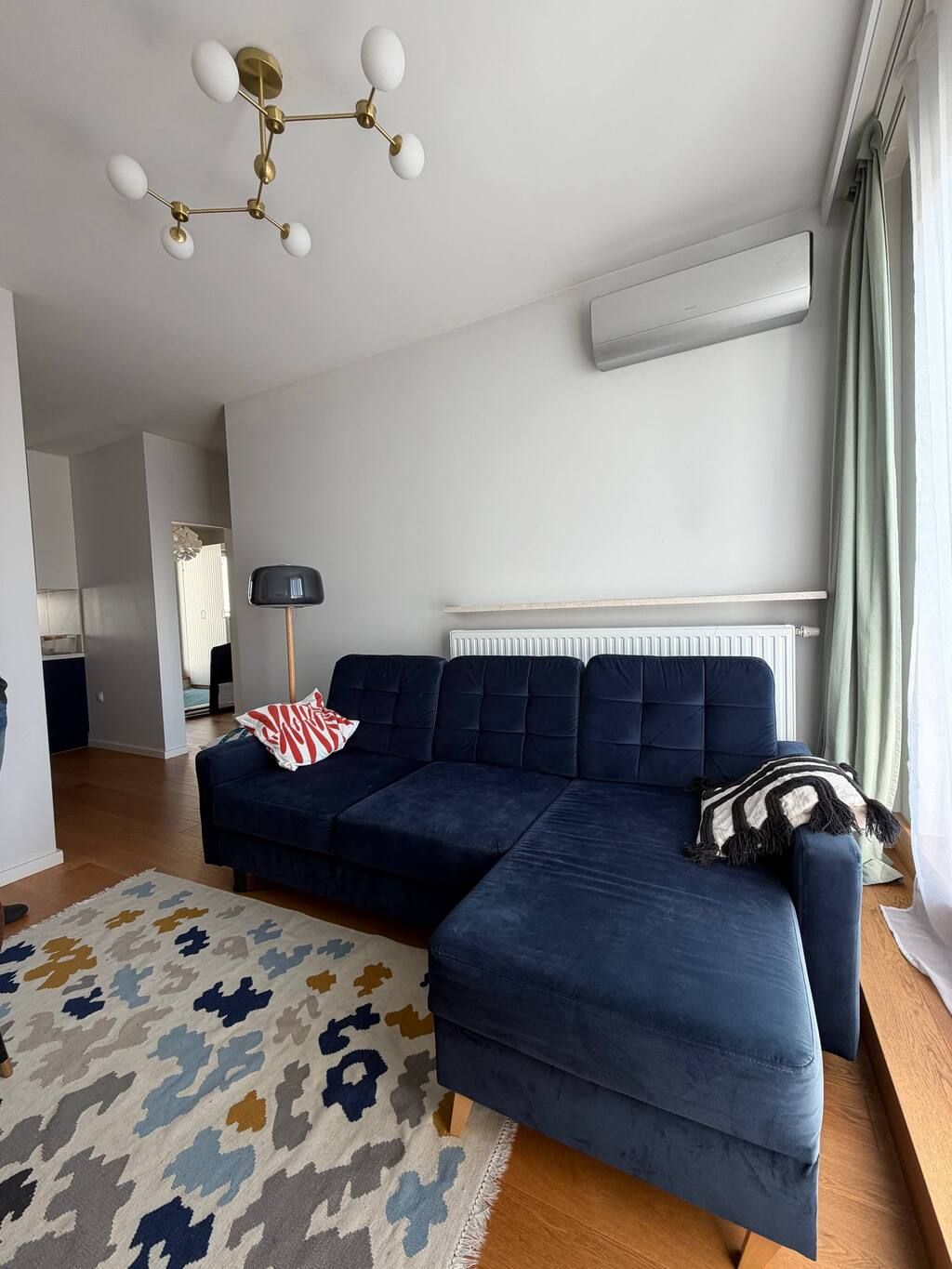 A modern living room with a sectional sofa, a patterned rug, a chandelier, and a radiator.