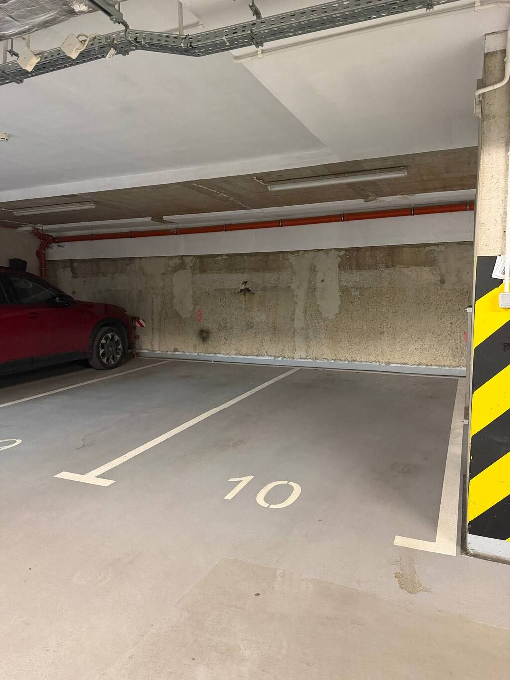A parking garage with concrete walls, a red car parked in one of the spaces, and a yellow and black striped safety sign on the wall.