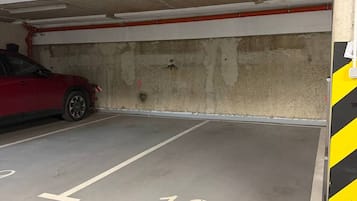 A parking garage with concrete walls, a red car parked in one of the spaces, and a yellow and black striped safety sign on the wall.