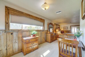 A room with wooden paneling, a large window with blinds, a wooden dresser, a mirror, and a table with a chair.