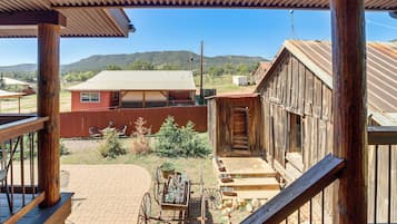 A rustic wooden building with a porch, a covered outdoor seating area, and a view of a distant house and mountains.
