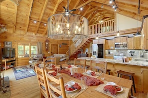 A spacious wooden cabin interior with a dining area, kitchen, and a staircase leading to an upper level. - Mtn Views From Every Deck Without The Mtn Drive! (Maggie Valley)