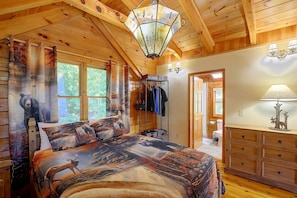 A cabin interior with a bed featuring a forest-themed bedding, a wooden dresser, and a hallway leading to a bathroom. - Mtn Views From Every Deck Without The Mtn Drive! (Maggie Valley)