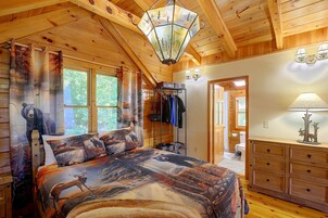 A cabin interior with a bed featuring a forest-themed bedding, a wooden dresser, and a hallway leading to a bathroom.