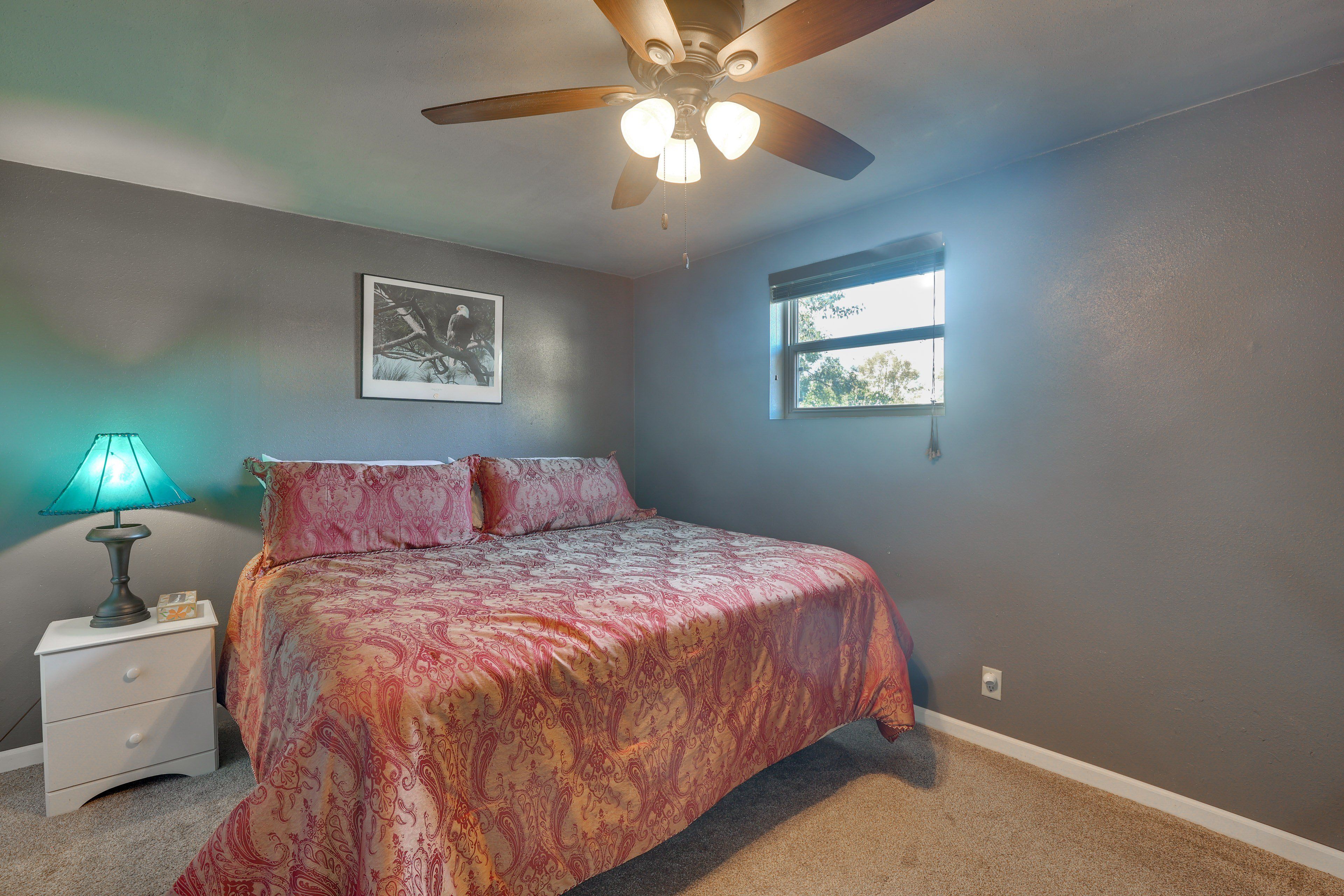 A bedroom with a bed, a nightstand, a ceiling fan, a window, and a wall-mounted picture.