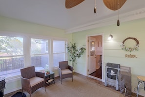 A room with wicker chairs, a potted plant, a dining table, and a small kitchen area with a refrigerator and a stool.