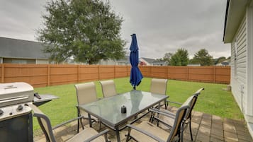 Un patio exterior con una mesa y sillas de vidrio, una parrilla y una sombrilla azul.