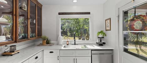 A modern kitchen with white cabinetry, a built-in dishwasher, and a window overlooking a garden.