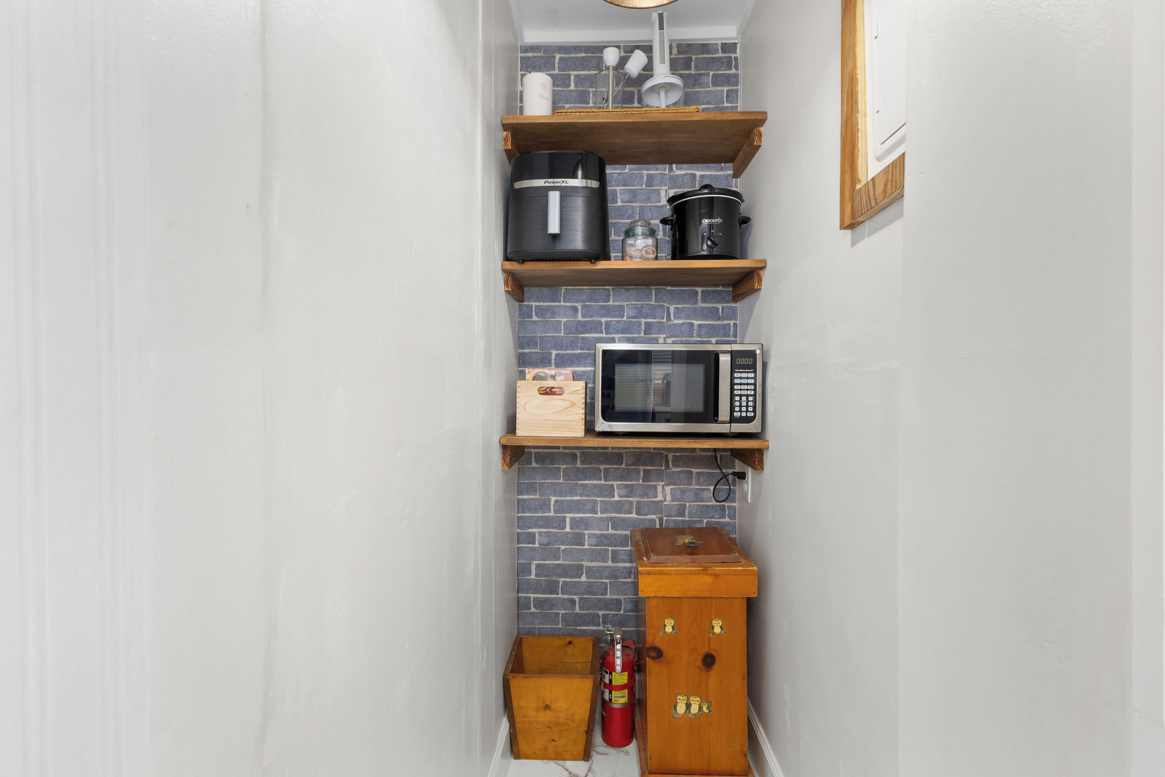 Une cuisine avec un mur en briques, des étagères en bois, un four à micro-ondes, une bouilloire et un extincteur.