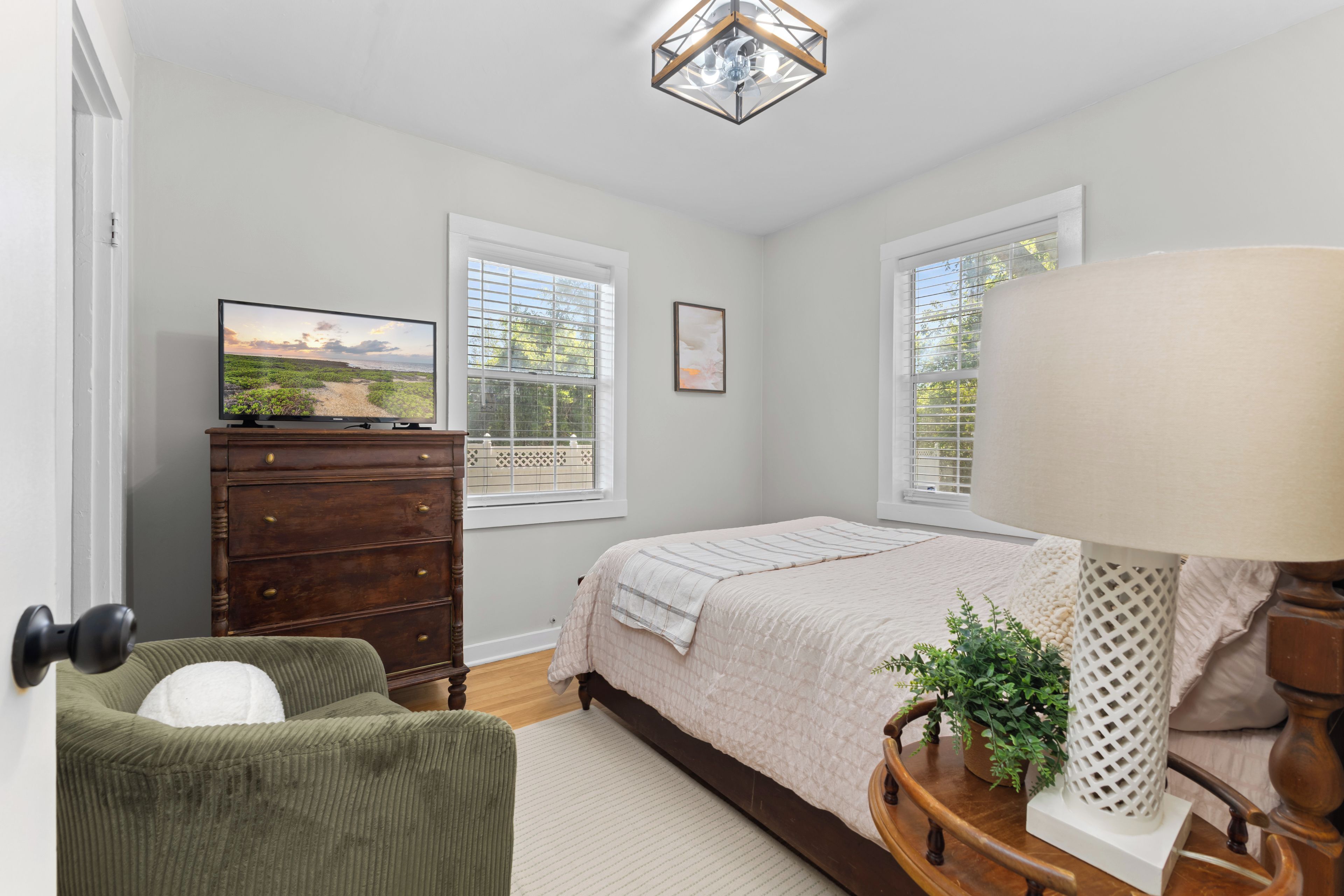 Un dormitorio con cama, cómoda, televisión, ventanas y lámpara.